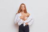 Pretty red-haired woman posing inside white studio, showing one naked boob with nipple sticker, wearing white shirt and black pants, beautiful body concept, copy space