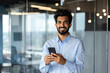 © Tetiana - Portrait of Indian young male student standing inside campus, office, university and using phone. Smiling and looking at the camera