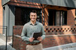 © LIGHTFIELD STUDIOS - confident real estate agent holding folder and looking at camera near modern house on street