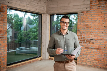 Wall Mural - joyful real estate agent in eyeglasses holding folder with pen and looking at camera in new house