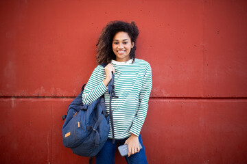 cheerful young african american woman standing by wall with school bag