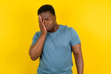 Tired overworked Young handsome man standing over yellow studio background has sleepy expression, gloomy look, covers face with hand, has eyes shut, gasps from tiredness, fatigue after party