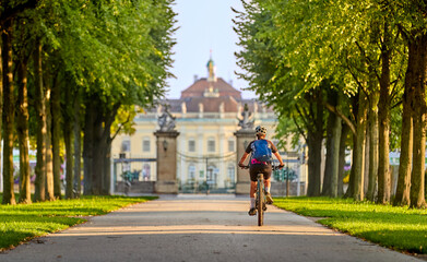 Wall Mural - attractive senior woman cycling with her electric mountain bike in an old avenue in Ludwigsburg, Baden-Wuerttemberg, Germany with famous Baroque castle in background
