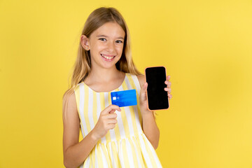 Photo of adorable Beautiful caucasian kid girl wearing yellow dress holding credit card and Smartphone. Reserved for online purchases