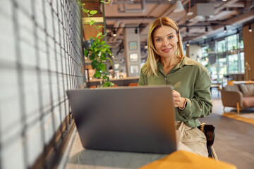 Wall Mural - Smiling woman sales manager working on laptop in cozy coworking and looking at camera