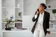 Attractive black business lady chatting with colleagues on cellphone, leaning on table in light office, free space