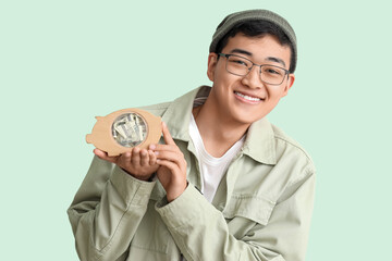 Poster - Young Asian man with piggy bank on green background