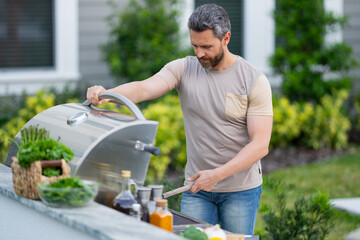 Wall Mural - Man at barbecue grill. Male cook preparing barbecue outdoors. Bbq meat, grill for picnic. Roasted on barbecue. Man preparing barbeque in the house yard. Barbecue and grill. Cook using barbeque tongs.