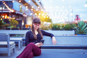 portrait beautiful young caucasian woman sitting on a bench outdoors looking at the cell phone smiling