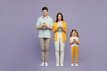 Wall Mural - Full body young fun parents mom dad with child kid daughter girl 6 years old wear blue yellow casual clothes hold in hand use mobile cell phone isolated on plain purple background. Family day concept.