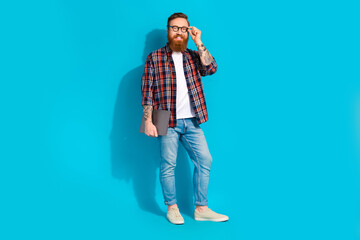 Full size photo of smart good mood man wear plaid shirt denim pants hold laptop touching glasses isolated on blue color background