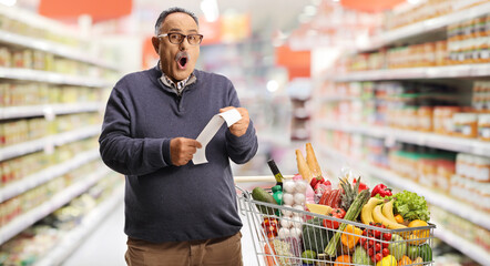 Wall Mural - Shocked mature man with a shopping cart holding a bill at a supermarket