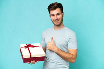 Young caucasian man holding a gift isolated on blue background with thumbs up because something good has happened