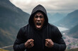 Anger African Man In Black Hoodie On Mountain Scenery Background . Сoncept Anger As A Tool For Survival, Representation Of African Men In Media, The Hoodie As A Sign Of Protection