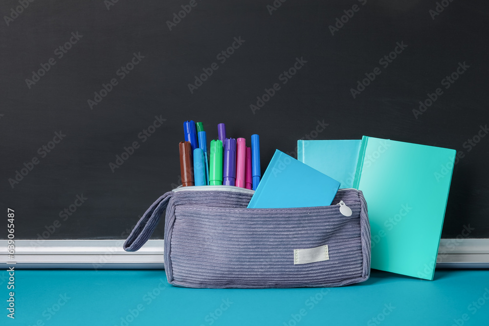 Pencil case with different school stationery on blue table near blackboard