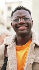 Wall Mural - Vertical portrait of young african american student with googles smiling and looking at camera standing at the high school, at the back ground a group of teenagers walking and laughing. Education