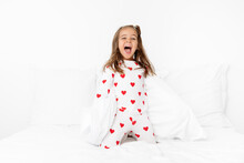 Excited Little Girl Wearing Pajama With Printed Hearts Kneeling On Bed With Mouth Open