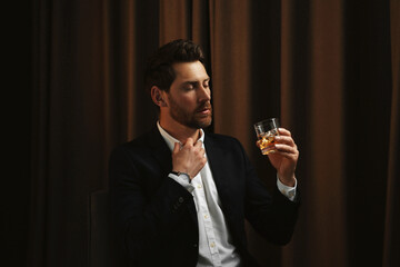 Canvas Print - Man in suit holding glass of whiskey with ice cubes on brown background