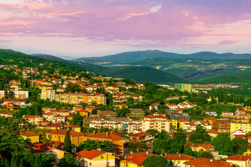 Wall Mural - panoramic landscape view of green summer city with yellow houses, green forest and mountains above and amazing hills and cloudy sky on background