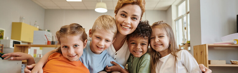 cheerful teacher and interracial kids looking at camera during lesson in montessori class, banner