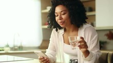 Charming Young Curly Woman Drinking Clean Water From A Glass While Scrolling Browsing On Smartphone Checking Email At Home Pretty Female Keep Health Balance Hydration During Day Indoors