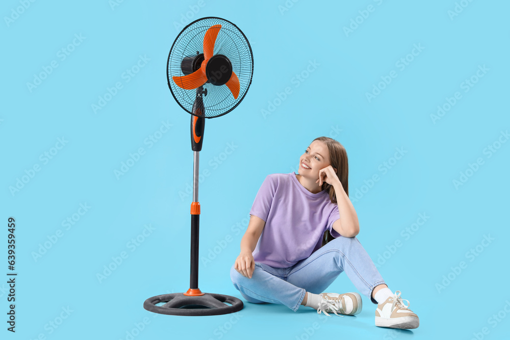 Young woman with electric fan sitting on blue background