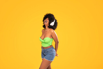 Adorable african american woman with white headphones laughing out loud, dancing, yellow vibrant studio background. Real people emotions