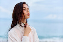 Portrait Of A Beautiful Pensive Woman With Tanned Skin In A White Beach Shirt With Wet Hair After Swimming On The Beach Ocean Sunset Light With Clouds