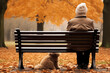 Back view of a person sitting on a park bench with a dog. The person is wearing a brown jacket and a beige beanie, orange leaves, autumn
