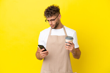 Poster - Restaurant waiter blonde man isolated on yellow background holding coffee to take away and a mobile