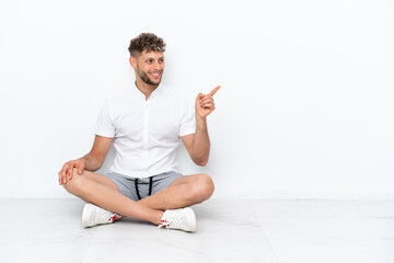 Canvas Print - Young blonde man sitting on the floor isolated on white background pointing finger to the side and presenting a product