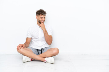 Wall Mural - Young blonde man sitting on the floor isolated on white background looking to the side