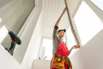 Wall Mural - Young man wearing overalls sealing cracks between window and trim using waterproof silicone caulk on the balcony.