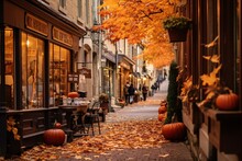 A Cobbled Street In A Historic Town. Burnt Orange And Deep Red Leaves Have Fallen And Scatter The Path. Shop Windows Display Pumpkin Arrangements And Autumn-themed Decor