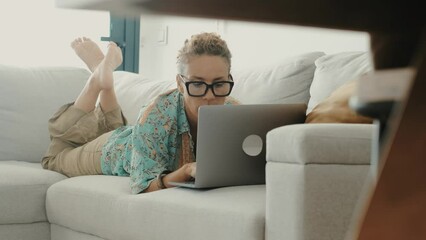 One adult woman using laptop laying comfortable on the sofa at home in casual clothes. Happy and easy lifestyle modern people working on computer and surfing the net. Smart work small business job