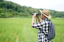 Asian Man Naturalist Wears Hat, Plaid Shirt, Backpack, Uses Binocular To Explore Nature At Paddy Field. Concept, Nature Exploration. Ecology Study. Pastime Activity, Lifestyle. Explore Environment.   
