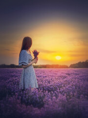 Side portrait of a young woman in dress stands in the purple lavender field looking at the beautiful sunset sky. Natural summer dusk scene.