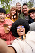 © CarlosBarquero - Vertical happy selfie of multi-ethnic group young friends looking laughing at camera. Portrait of multiracial students hugging and sitting in park. Generation z people photo outdoors spring day.