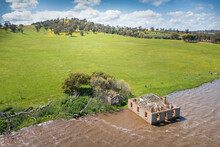Aerial View Of An Abandoned Historic Farm House Full Of Water On The Edge Of A Lake Below Green Hill