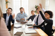 © fizkes - Motivated multiracial employees sit gather at desk in office raise hands participate in teambuilding activity together, diverse colleagues vote volunteer at team briefing or meeting, training concept