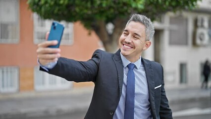 Wall Mural - Young hispanic man business worker smiling confident make selfie by smartphone at street