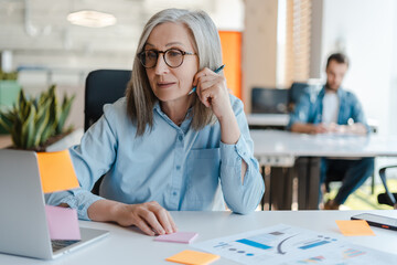 Smiling confident senior businesswoman using laptop, sticky notes looking at monitor in modern office. Gray haired manager wearing stylish eyeglasses  sitting at workplace. Scrum, agile concept 