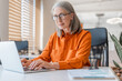 © Maria Vitkovska - Confident smiling mature businesswoman wearing stylish eyeglasses, orange shirt using laptop working online in modern office. Manager, writer planning project, typing on keyboard sitting at workplace