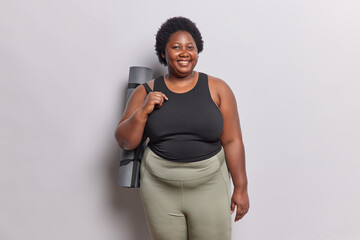 Wall Mural - Body positive dark skinned woman dressed in black t shirt and leggings carries rolled rubber karemat going to have fitness training with coach poses against white background practices yoga every day