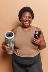 Wall Mural - Body positive dark skinned woman holds foam roller smartphone going to exercise and make massage dressed in tracksuit smiles gladfully isolated over brown background exercising muscles before training