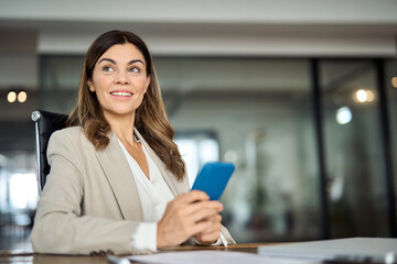 Smiling mature middle aged 40 years old professional business woman executive holding smartphone using cell phone mobile apps looking away working on cellphone sitting at office desk. Copy space