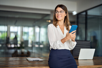 Wall Mural - Happy mature business woman executive holding cell phone looking away in office. Smiling mid aged 40s professional businesswoman manager entrepreneur using cellphone working on smartphone. Copy space