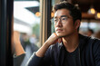 © Justlight - A thoughtful Asian man sits at a cafe watching people pass by his window. His dark eyes peer through his glasses as he contemplates his identity. The light shadows the contours of