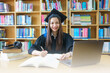 © EduLife Photos - An asian girl student in university graduate gown with books, laptop studies happily in library.