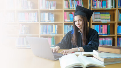 Wall Mural - An asian girl student in university graduate gown with books, laptop studies happily in library.